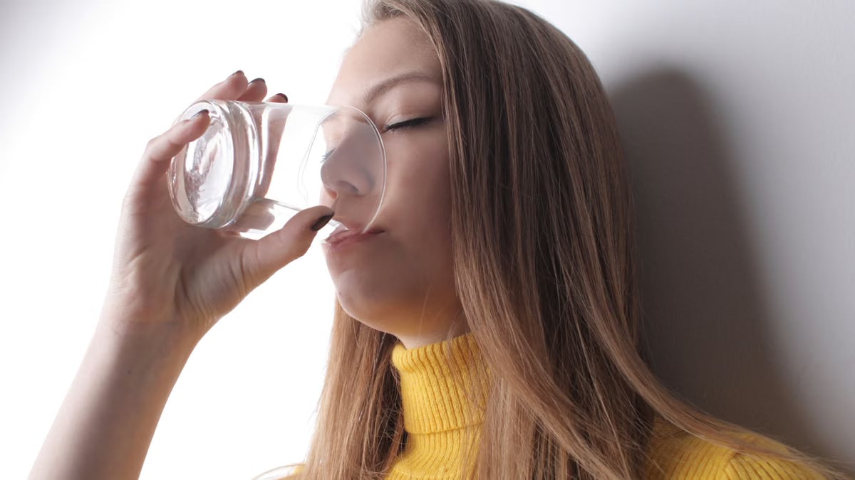 Woman in a yellow sweater drinks water from a transparent glass.