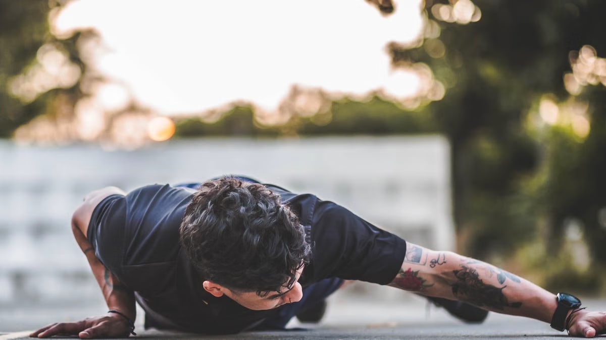 Tattooed man doing a side plank exercise outdoors in a sunset environment.